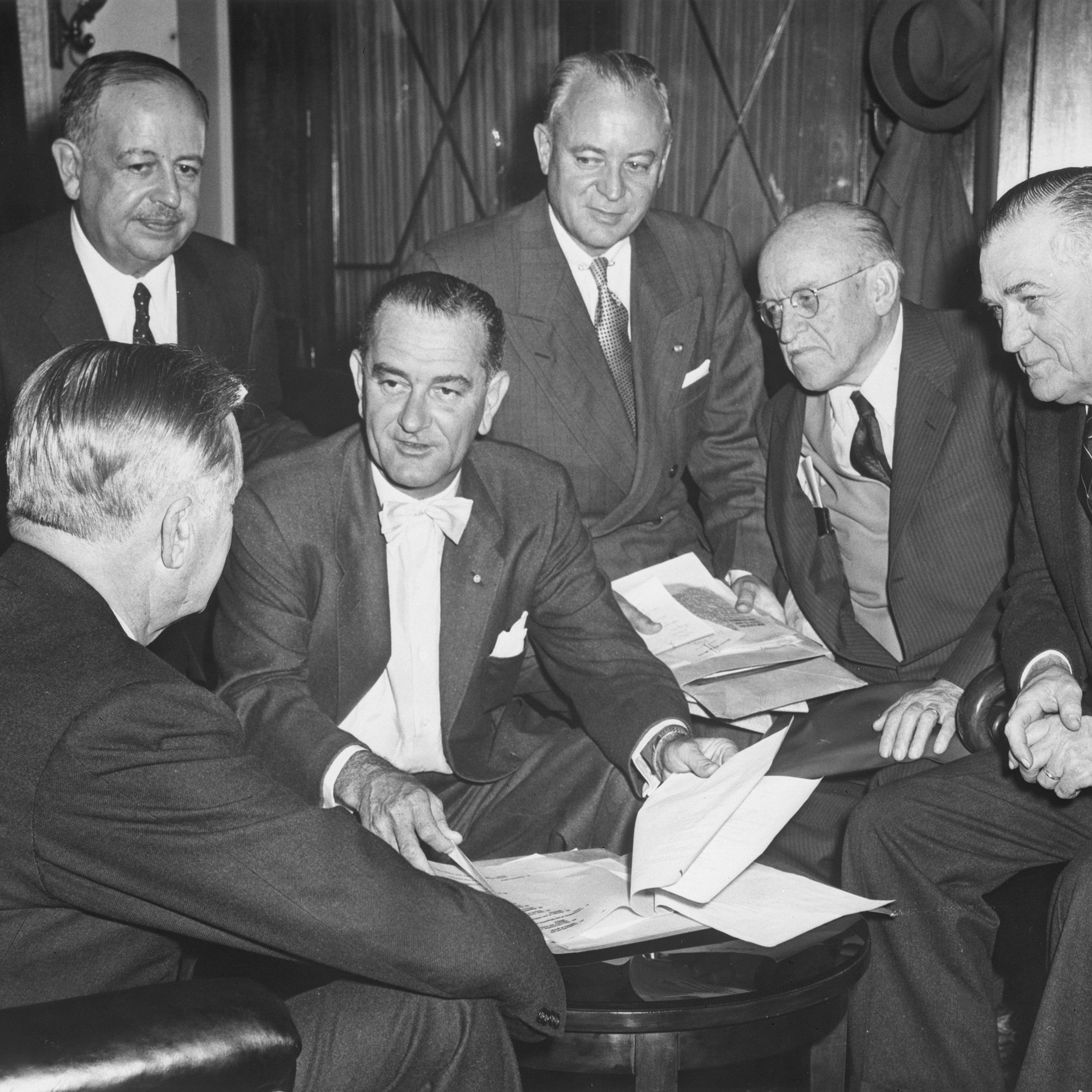 Six men in suits in conversation. LBJ (center) looks through some papers.